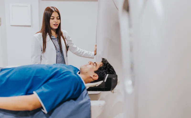 Baş tutucuya yerleştirilmiş hasta görüntüleme cihazının masasında yatarken yanında duran kadın doktor